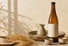 生成り色の空間に日本酒瓶と燗酒器、稲穂を配した写真